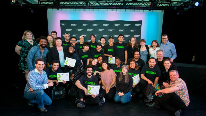 A group of TWDP graduates posing in a black box theatre in from of a black and white RTC step and repeat.