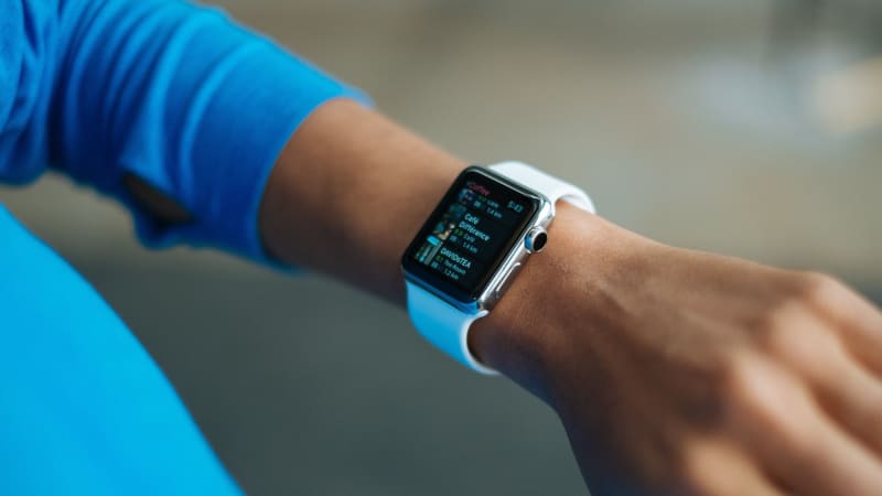 The wrist and lower arm of a medium-skinned person wearing a bright blue microfiber shirt and an Apple Watch. The watch face displays listings for local coffee shops.