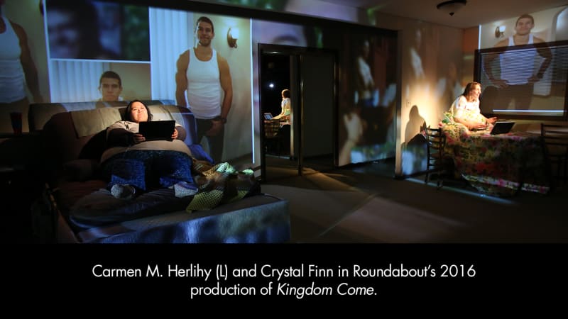 Producton photo from Kingdom Come. Two women on either side of a room sit up in bed on their laptops. Images of musclar men are projected on the walls.