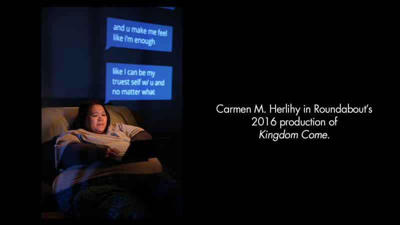 Production image from KINGDOM COME. A heavy woman with light-medium skin and dark hair types on a laptop, illuminated by its light. Text messages are projected onto the wall behind her.