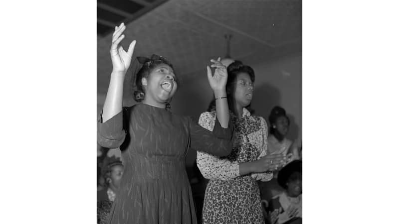 A dark-skinned woman in a dress and small hat sings, raising her hands in the air. A second dark-skinned woman in glasses, patterned dress, and hat sings with eyes closed.