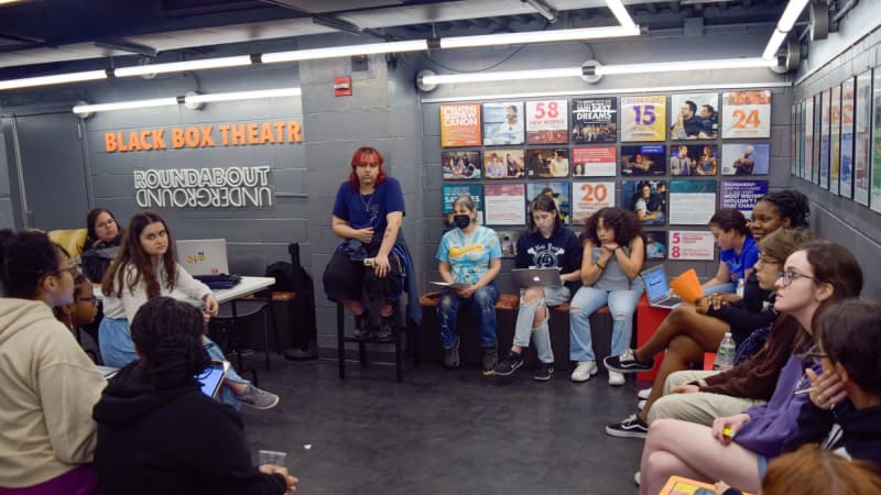 A large group of young people of different skin tones sits in a circle in the lobby of a black box theatre.