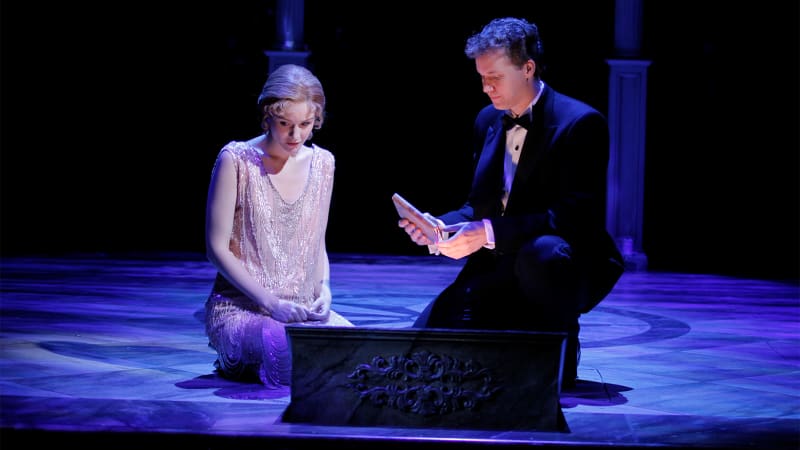 A young, light-skinned woman in a beaded evening dress kneels beside a light-skinned man in a tuxedo. The man holds a book. 