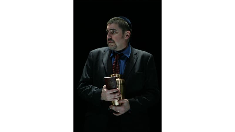 A light-skinned man with dark hair and a goatee wears a dark suit and yarmulke. He holds an urn and a prayer book.