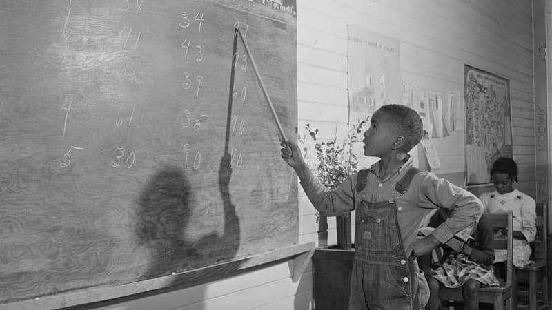 A dark-skinned young boy in denim overalls and a button-up shirt stands with his left hand on his hip, his right hand holding a pointer. He points to a number written on a chalkboard.