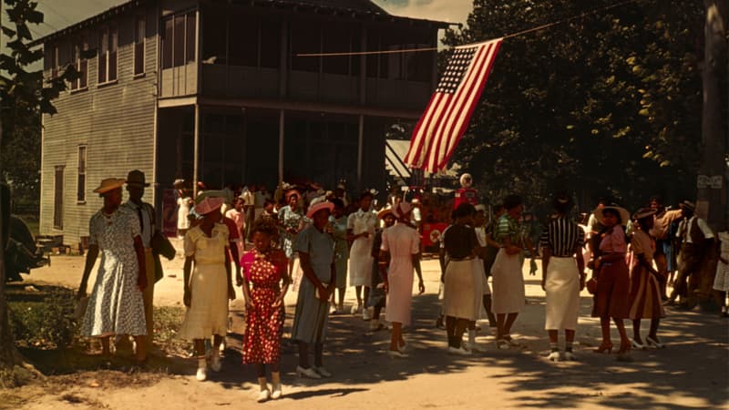  Dark-skinned women in colorful, special-occasion dresses and skirts congregate near a large American flag on a sunny day. 