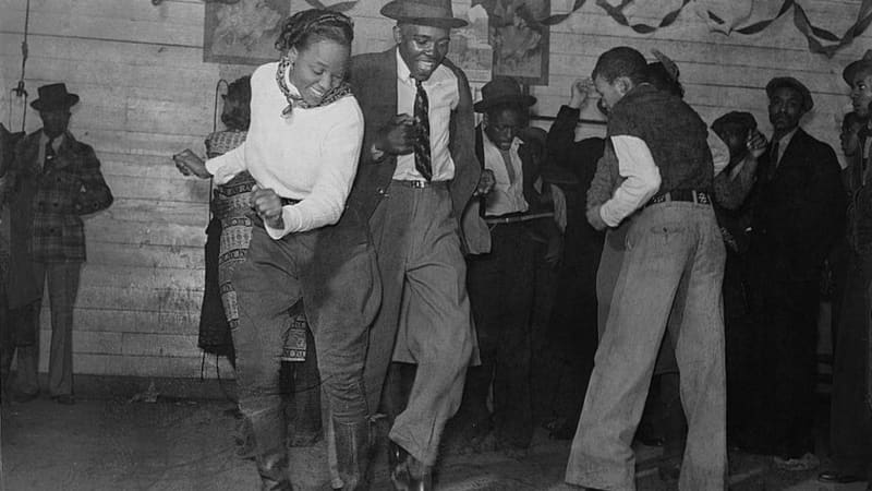 A dark-skinned woman in casual shirt, pants, and boots dances with a dark-skinned man in a suit, tie, and hat. Behind them, others dance. Streamers hang from the walls.