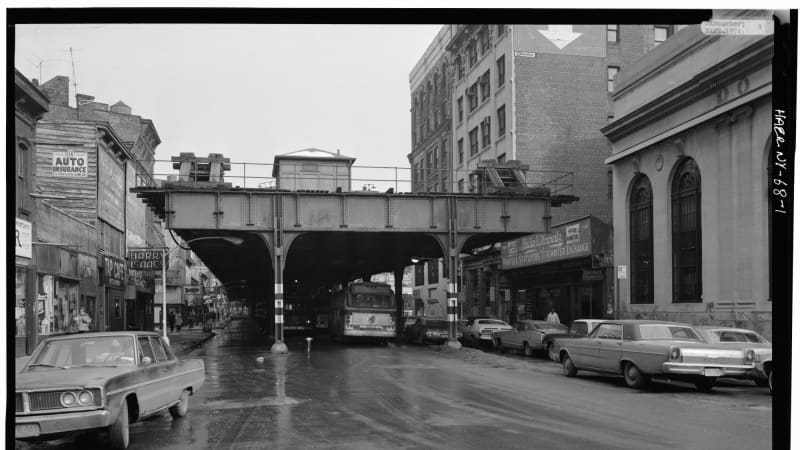 A wide city street disappears under elevated rail tracks. A mix of one-to-six story buildings, some of them wooden, stand on both sides of the street.