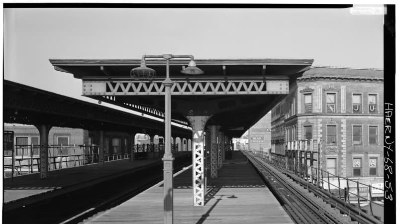 An empty elevated subway platform. Snow blankets the uncovered section of platform. The tracks are hemmed in by mid-rise apartment buildings.