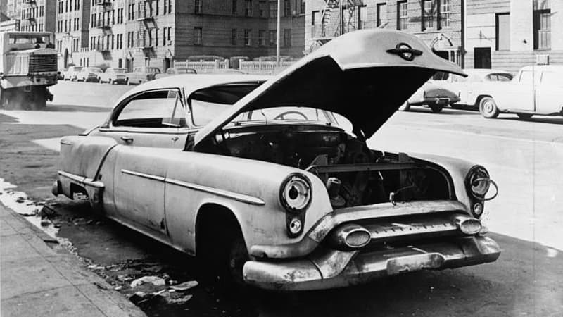 A 1950s car with its hood open, stripped of tires and parts, sits on a wide street. Across the street rise six-story apartment buildings with fire escapes.