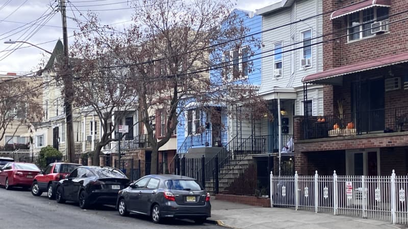 A row of well-kept two-and-three story houses. Their walls touch. They are red brick, white siding, blue siding, and beige siding. Cars are parked on the street in front of them.