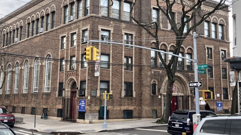 A four-story brown brick school building, with tan moldings around the windows and a red front door. A bare tree grows out of the sidewalk in front, and cars are parked on the streets around it.