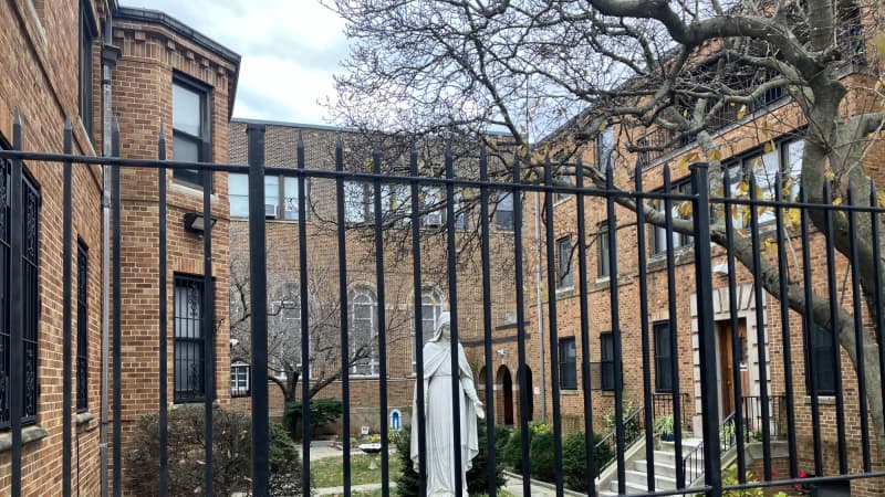 : The courtyard of an orange-brick, U-shaped building seen through a black wrought iron fence. The courtyard is grass, with beds of shrubs and flowers and features a white statue of Mary. 