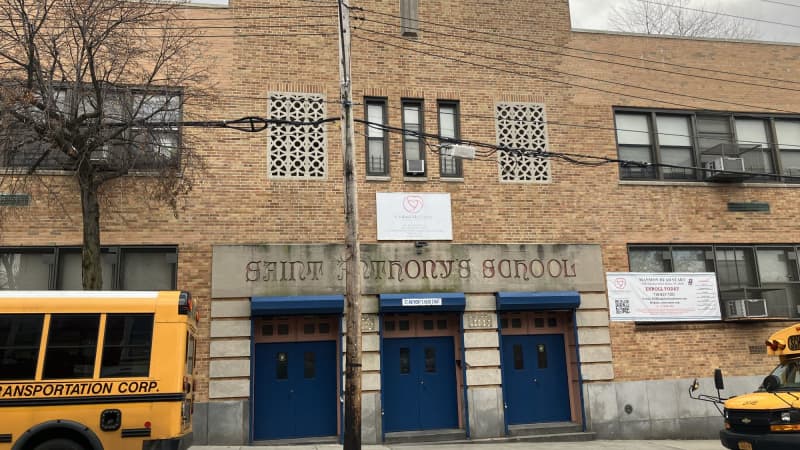 The front of a school building: the center of the building is brown brick, rising three stories, with a cross in the top center. The wings are two stories and orange brick. Three sets of blue doors si