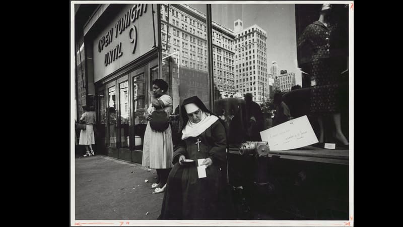 A store front. An older, light-skinned nun in her habit sits in front, looking at something in her lap. Other women stand nearby.