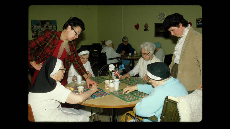 Four light-skinned elderly women sit at a table with bingo cards spread in front of them. Three wear habits with headpieces. Two younger light-skinned women in street clothes stand near them, helping.