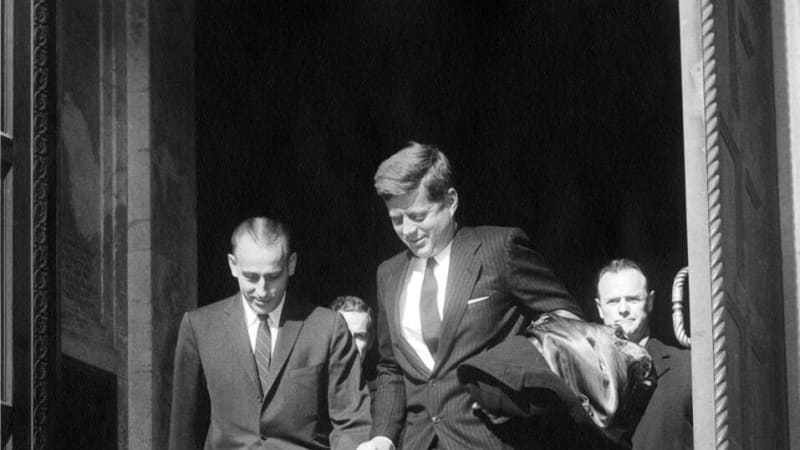 Two light-skinned men in dark suits exit a tall door into a sunny day.