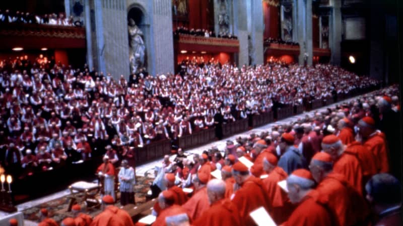 Hundreds of men in red-and-white cassocks and hats stand inside a large church. In the center aisle, a similarly-dressed man stands behind a gold table.