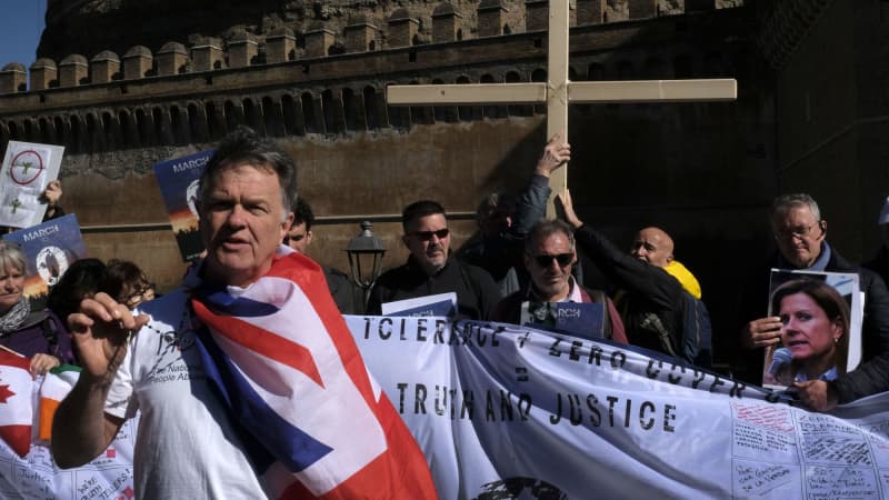 A light-skinned, middle-aged man wearing a blue, red, and white flag around his neck holds a rosary and stands in front of a row of protestors who hold a banner reading “Zero Tolerance and Zero Cover-