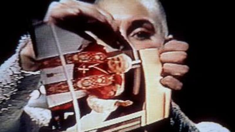 A light-skinned woman with a shaved head holds a photo of a man in ornate red-and-white clerical dress in front of her face and rips it.