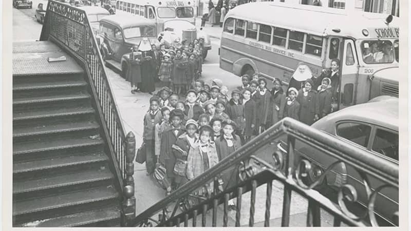 A crowd of children with dark and medium skin tones exit school buses, accompanied by two light-skinned nuns in dark tunics and white headpieces.