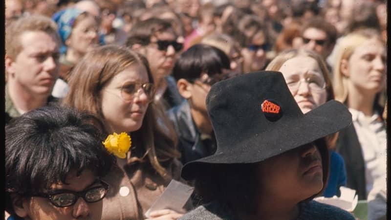 Two medium-dark skinned women at a protest. The woman on the left has a yellow flower in her hair. The woman on the right wears a wide-brimmed hat. Both wear many buttons.