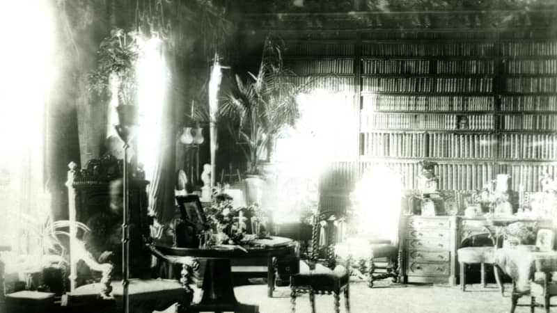  A high-ceilinged library,t he back wall is covered in books. Two tall windows let light in from the left. A faint, ghostly figure of a person sits in a chair.