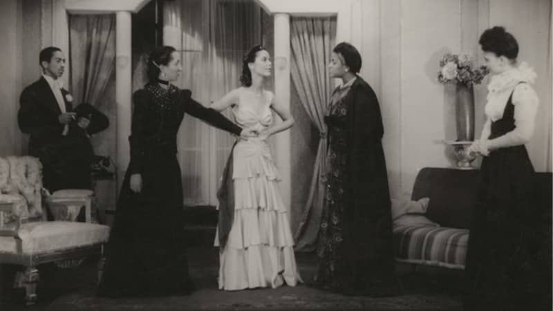 A young Black woman in a light dress with hands on her hips speaks with two older Black women in dark dresses. A Black man in a tuxedo and a Black woman in a frilly shirt and black jumper look on.  