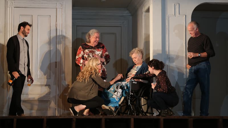  A young, bearded man holding a wine bottle looks on as two young women, a middle-aged woman, and a middle-aged man surround an elderly, light-skinned woman in a wheelchair.  