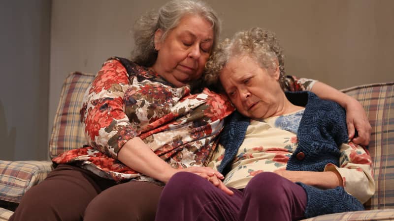 A light-skinned, middle-aged woman in a floral shirt has her arm around an older, light-skinned woman wearing a blue knit vest. The older woman rests her head on the younger woman’s shoulder.  