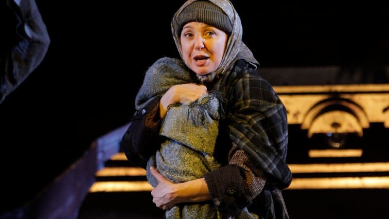 A light-skinned woman in a heavy, dark dress, shawl, and winter hat clutches a blanket-wrapped baby. 