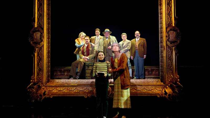 An older, light-skinned woman in house dress and cardigan stands next to a young girl in a striped shirt. Behind them a large, ornate picture frame in which six light-skinned adults stand posed.  