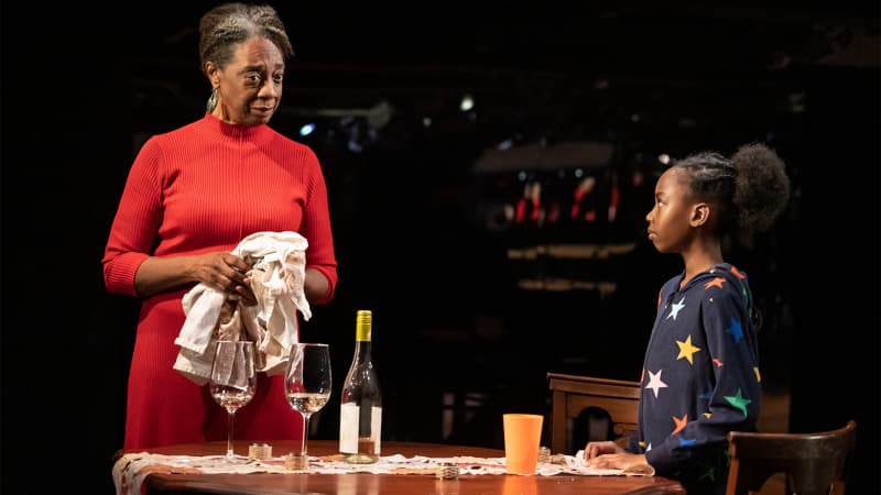 An older, dark-skinned woman in a red dress stands behind a half-cleared dinner table and speaks to a young, dark-skinned girl wearing a navy-blue hoodie with colorful stars on it. 