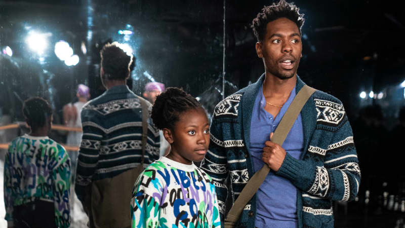 A young, dark-skinned girl in a sweatshirt and track pants stands next to a dark-skinned man in sneakers, a colorful cardigan, and a messenger bag. They are reflected in a large mirror behind them. 