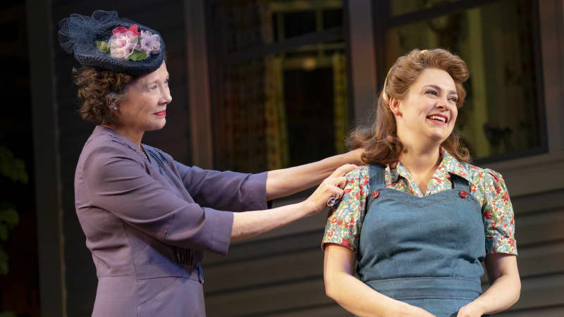 A older woman with a fancy Sunday hat and simple purple dress places her hands on the shoulders of a younger woman wearing a denim jumpsuit over a floral short sleeved shirt.