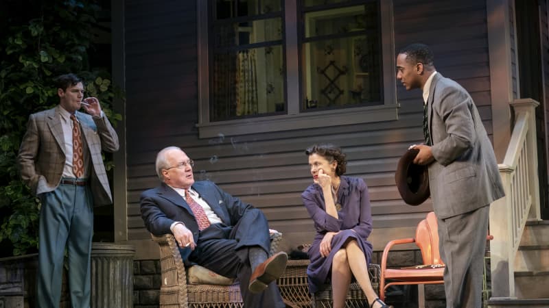 An old man and woman sit on wicker furnature outside of a home. A man smokes a cigarette behind them and a Black man holding a fedora speaks to them.