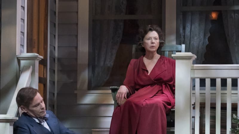 An older woman with brown curls and a red robe sits on a green rocking chair on the porch of a home. A man in a suit sits on the steps below her.