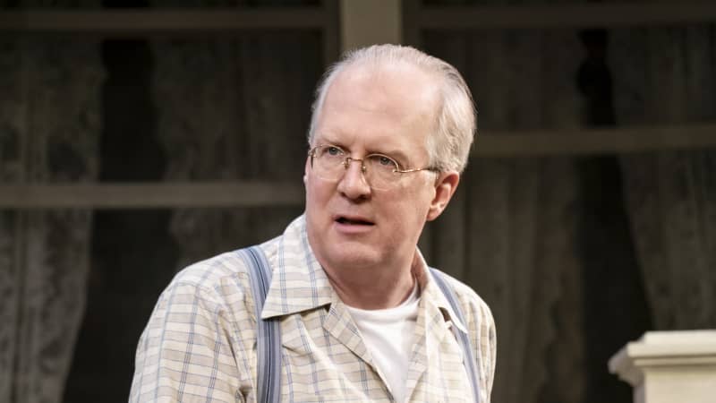 An older man with thin glasses, a checkered collared shirt, blue pants, and overalls stands in front of a home.