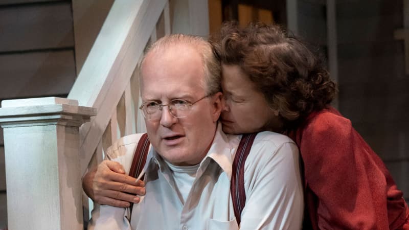 An old woman with brown curly hair in a red robe sits on the stairs to a house and burrows her face into the neck of an old man in suspenders and glasses who sits next to her.