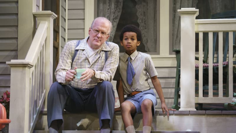 An old man in suspenders sits next to a young boy on the front steps of a home.