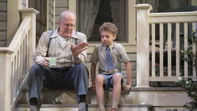 An old man in suspenders sits next to a young boy wearing a collared shirt and tie on the front steps of a home. The man holds a coffee cup and the kid looks nervous.