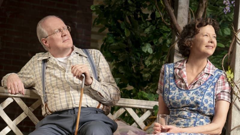 An old man in suspenders and a cane and an old woman wearing a blue floral apron over a pink checkered dress sit together on a garden bench.