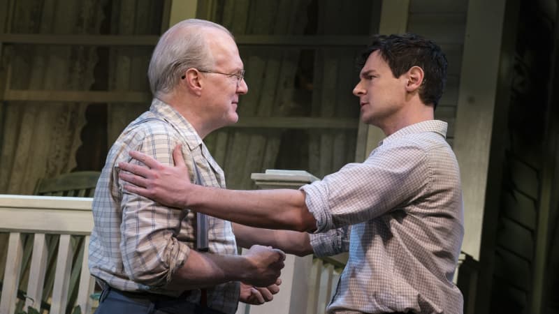 A man grabs the shoulders of an old man in suspenders who smiles at him outside a porch.