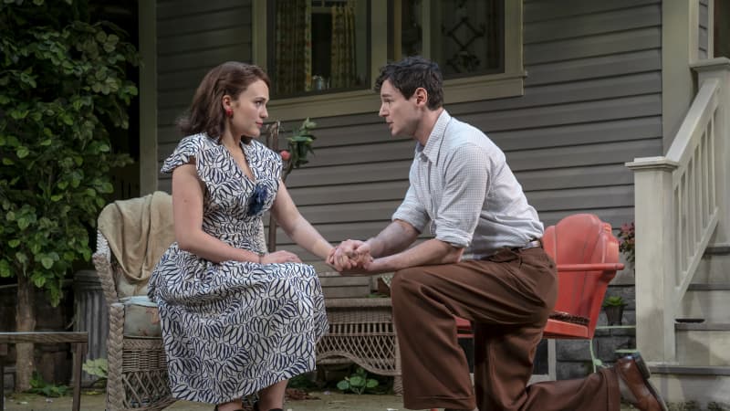 A woman in a white and black patterened dress sits in an outdoor chair while a man kneels down next to her and clasps his hands around hers.