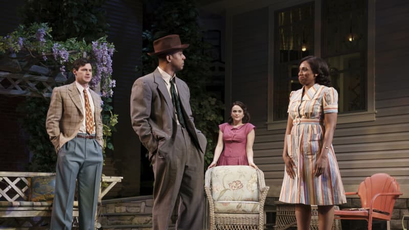 A Black man and woman speak with each other in the front lawn of a home while a white man and woman watch them in the background.