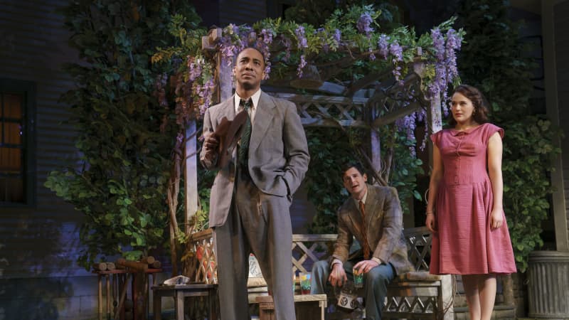 A Black man in a gray suit holds a brown fedora hat and stands in front of a garden bench and gazebo. A white man sits on the bench behind him, and a woman in a pink dress stands to the side.