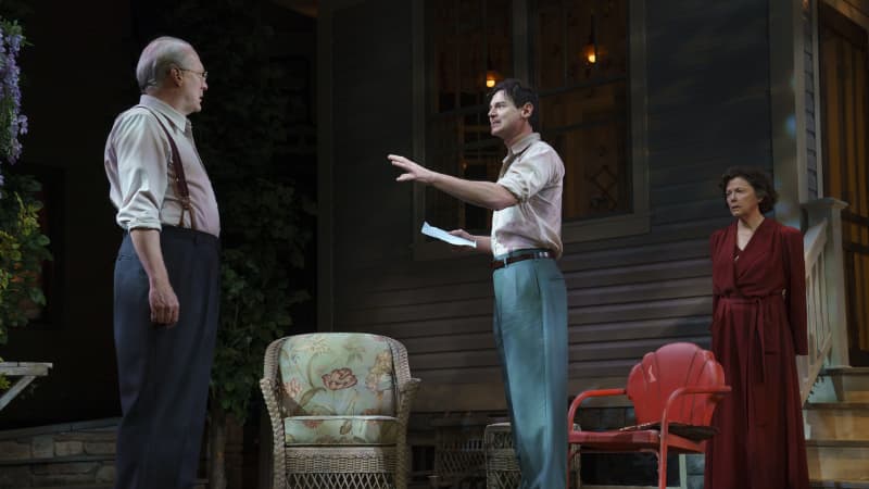 An man holding a piece of paper speaks with an arm outstretched to an old man on the front lawn of a home. An older woman in a red robe stands behind them with an upset expression.