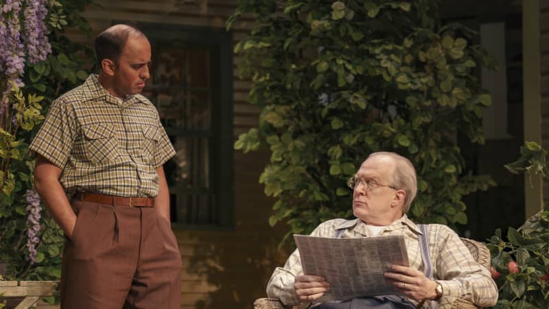 An older man sits in a wicker chair reading a newspaper and loops up at another man standing with his hands in his pocket. A lot of plants an greenery are in the background.