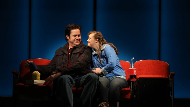 A man and woman on a date at a movie theater. The woman has Down syndrome and long brown hair. She rests her chin on the shoulder of the man, who smiles at her.