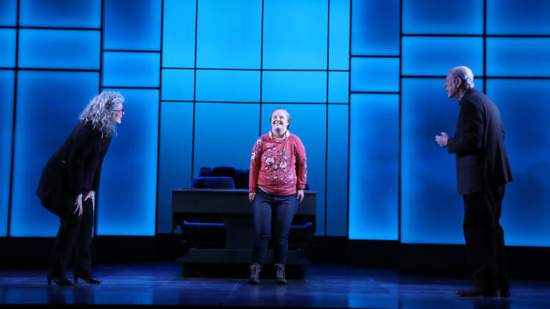 On a stage with bright blue paneling, a woman with Down syndrome smiles and wears a pink floral sweater. On either side of her are an older man and woman who speak to her.